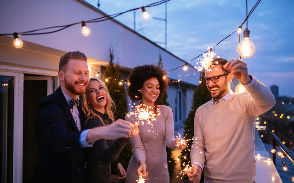 Vier Freunde mit leuchtenden Wunderkerzen in den Händen zu einer Party auf einer Dachterrasse in Abenddämmerung und mit Lichterketten