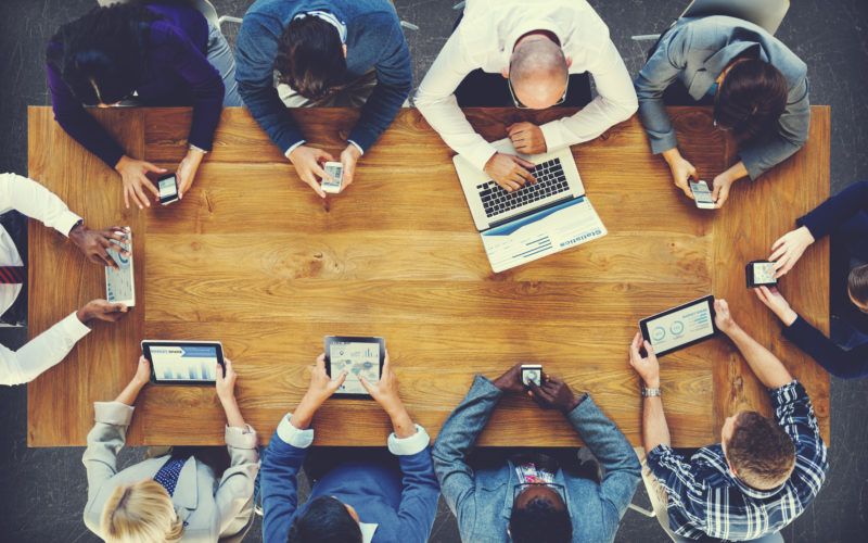 Menschen mit Laptops, Tablets und Smartphones sitzen um einen Tisch herum, Blick von oben