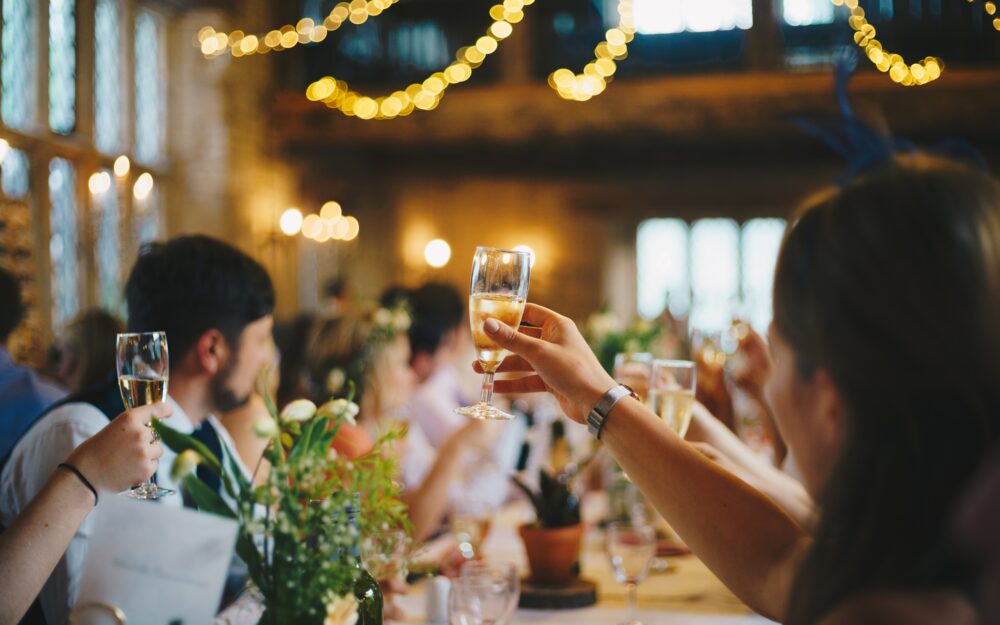 Festliches Anstoßen mit Champagner in einer festlichen Location, geschmückt mit Blumensträußen auf den Tischen und Lichterketten