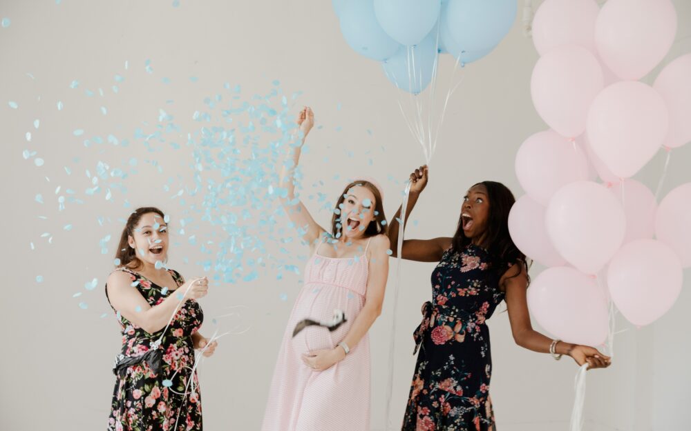Drei Frauen sind auf einer Babyshower-Party, zwei halten Ballons in Rosa und in Blau, der eine Ballon ist zerplatzt und enthüllt, dass das Baby ein Junge wird