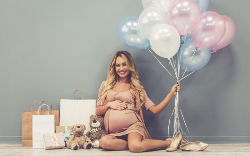 Schwangere, glückliche Frau sitzt auf dem Boden, hält Luftballons in der Hand, neben ihr stehen Geschenktüten