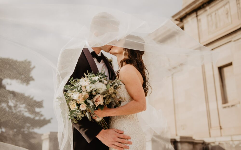 pexels-emma-bauso-2253870 Frisch verheiratetes Brautpaar küsst sich mit Blumenstrauß in der Hand