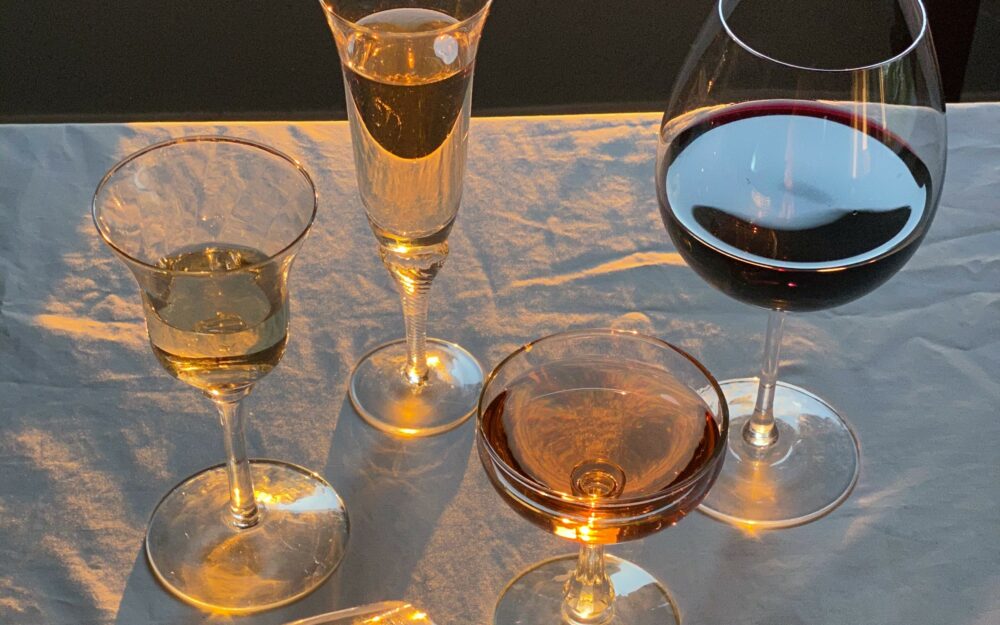Vier verschiedene gefüllte Weingläser stehen auf einer weißen Tischdecke und werden von Sonnenlicht betrahlt