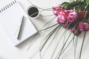 Auf einer weißen Unterlage liegen ein Notizbuch mit Stift, eine Kaffeetasse und ein Blumenstrauß
