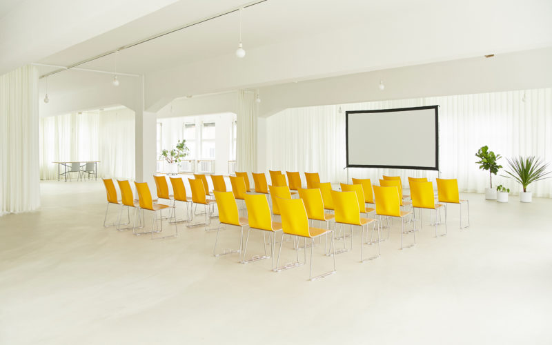 Weißer Boden und weiße Wände mit gelben Stuhlreihen und Beamer, Eventlocations für bis zu 200 Personen