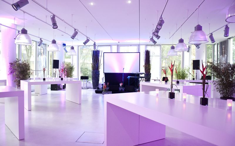 Heller moderner Veranstaltungsraum mit Glaswänden, weißen Tischen und einer kleinen Bühne