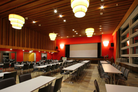 Großer Eventsaal mit einer Bühne, Tischen, Stühlen und rot bemalten Wänden
