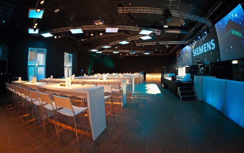 Große Eventhalle mit einer Bühne, weißen Tischen und Stühlen