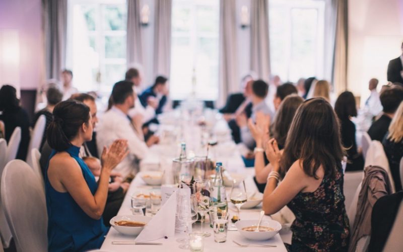 Lange Tischtafel mit weißer Tischdecke, Essen und klatschenden Menschen die daran sitzen, Traumhafte Firmenfeier in Bonn