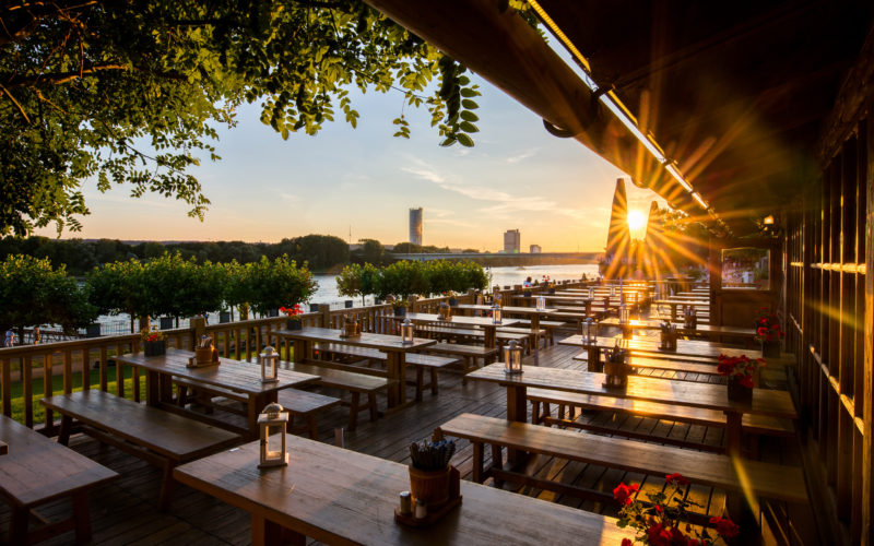 Restaurant Terrasse mit Blick über einen Fluss bei Sonnenuntergang. Rustikales Design mit Holztischen und Holzbänken