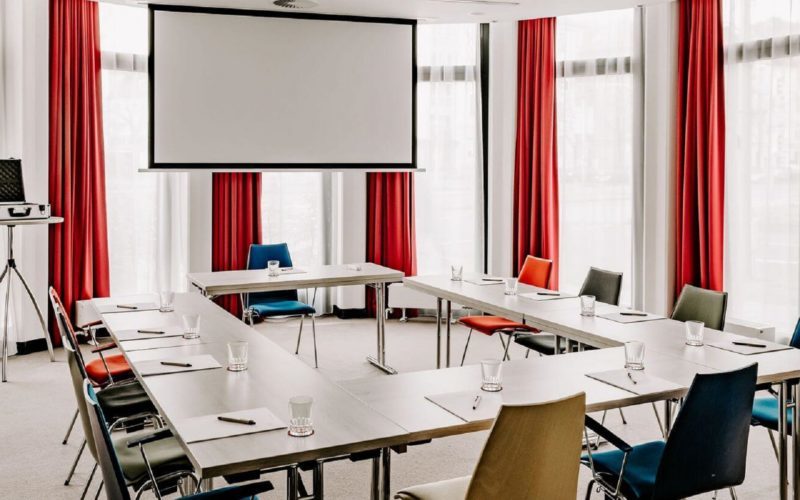 Konferenzraum mit roten Vorhängen, Tischen, Stühlen und einer Leinwand; Meetingraum in München