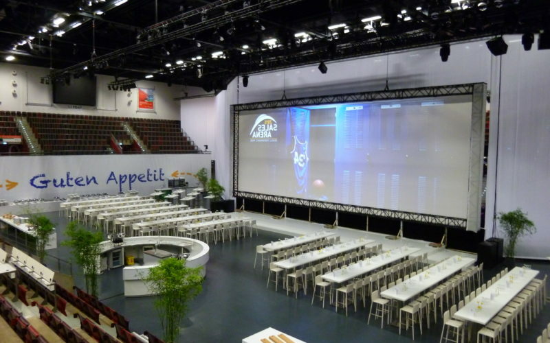Große Eventhalle mit vielen Tischen und Stühlenund mit großem Bildschirm