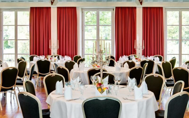 Eleganter Saal mit runden, weiß gedeckten Tischen, Stühlen und roten Vorhängen; Silvesterparty in Bamberg