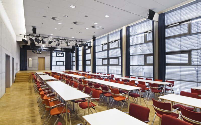 Moderne Eventlocation mit langen Tischen, roten Stühlen und einer Bühne; Firmenfeier in Kerpen