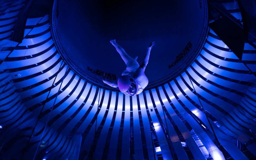 Ein dunkelblau beleuchteter Windtunnel mit einer skydivenden Person, aus einem Winkel von unten fotografiert